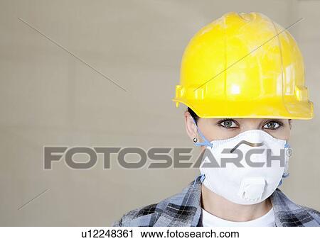 Stock Image - Portrait of female worker wearing dust mask and hardhat over colored background. Fotosearch