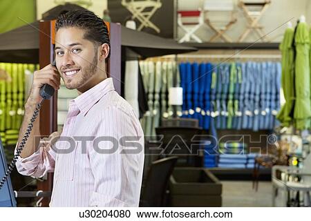 Stock Image - Portrait of handsome young employee in store listening to telephone receiver. Fotosearch