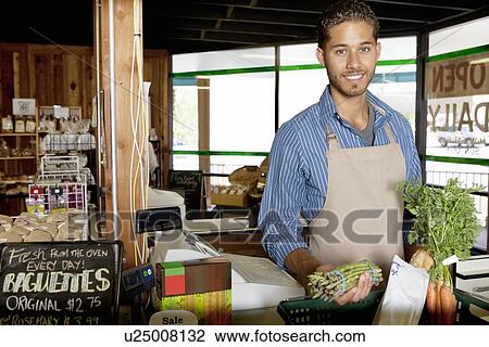 Stock Image - Portrait of handsome young store clerk holding vegetable in supermarket. Fotosearch