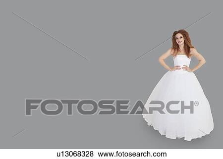 Stock Photo - Portrait of happy brunette in wedding dress with hands on hips standing over gray background. Fotosearch