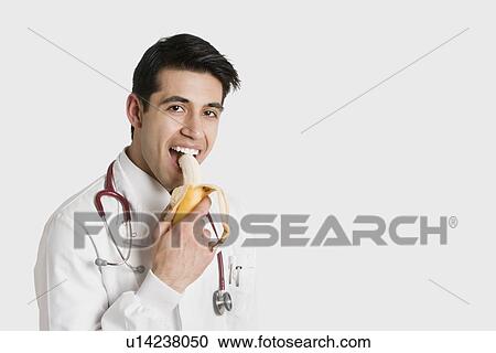 Stock Image - Portrait of Indian male doctor eating banana over white background. Fotosearch