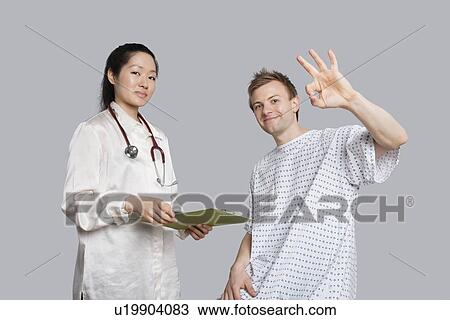 Stock Image - Portrait of patient gesturing okay with doctor holding a clipboard. Fotosearch