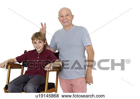 Portrait of senior man gesturing peace sign while pre-teen boy sitting on director's chair over white background View Large Photo Image Stock Photo - Portrait of senior man gesturing peace sign while pre-teen boy sitting on director's chair over white background. Fotosearch