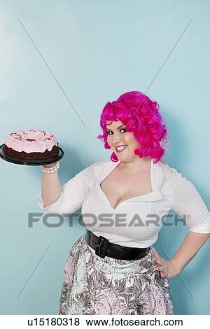 Portrait of young woman standing with hands on hips holding cake over colored background View Large Photo Image Stock Photo - Portrait of young woman standing with hands on hips holding cake over colored background. Fotosearch