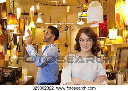 Stock Image - Portrait of young woman with arms crossed while man looking at price tag in background in lights store. Fotosearch