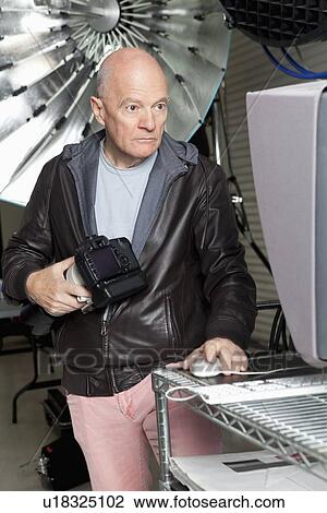 Stock Image - Senior photographer with camera using computer in studio. Fotosearch