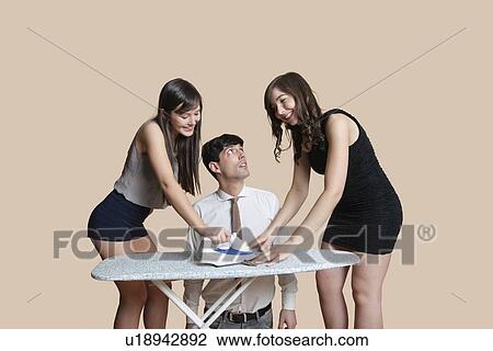 Stock Image - Shocked man looking at young females ironing tie over colored background. Fotosearch