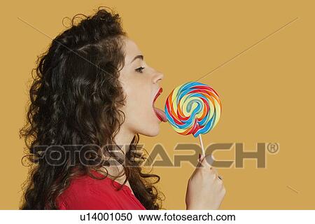 Side view of a young woman licking lollipop over colored background View Large Photo Image Stock Image - Side view of a young woman licking lollipop over colored background. Fotosearch