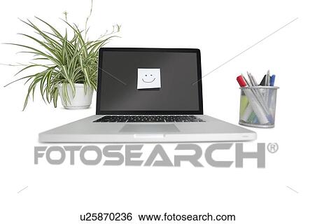 Stock Photograph - Smiley face sticky note on laptop with office supplies and pot plant. Fotosearch
