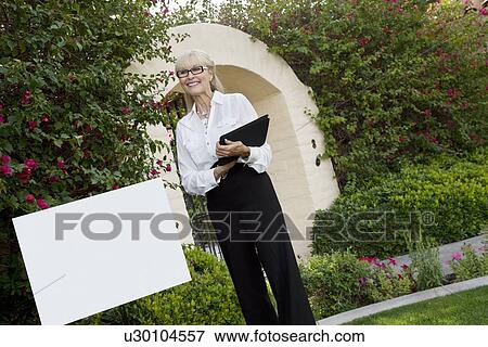 Tilt shot of cheerful senior agent standing by sign board with clipboard View Large Photo Image Stock Photo - Tilt shot of cheerful senior agent standing by sign board with clipboard. Fotosearch