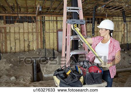Stock Image - Young architect looking at measuring tape at construction site. Fotosearch