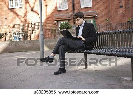 Young businessman using laptop while sitting on bench View Large Photo Image Stock Photography - Young businessman using laptop while sitting on bench. Fotosearch