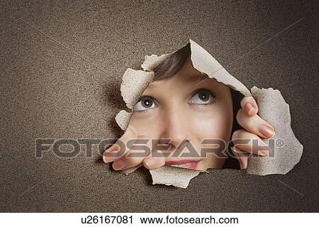 Stock Image - Young Caucasian woman peeking from ripped paper hole. Fotosearch