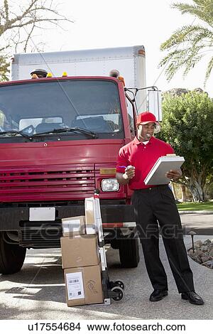 Young delivery man looking at delivery list on clipboard with truck in background View Large Photo Image Picture - Young delivery man looking at delivery list on clipboard with truck in background. Fotosearch