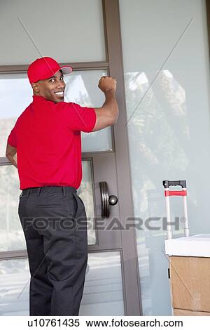 Stock Photography - Young delivery man with packages looking back while knocking on door. Fotosearch