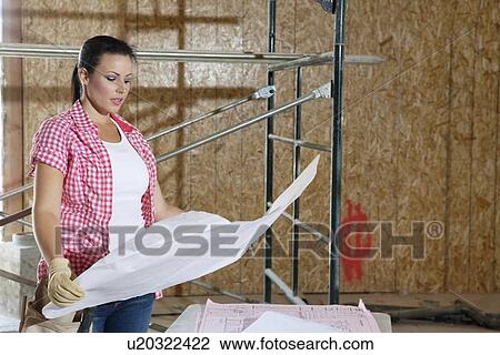 Stock Image - Young female contractor looking at building plans with scaffold in background. Fotosearch