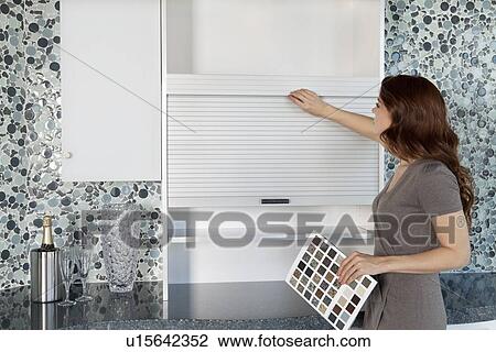 Stock Image - Young woman looking at cabinets in contemporary kitchen while holding color samples. Fotosearch