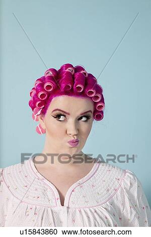 Stock Image - Young woman making faces with pink hair and curlers over colored background. Fotosearch