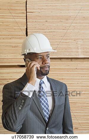 Stock Photo - African American male engineer on phone call with stacked wooden planks in background. Fotosearch