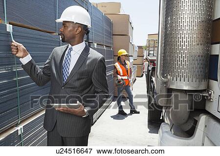 Stock Photo - African American male engineer inspecting planks while female worker standing by flatbed truck. Fotosearch
