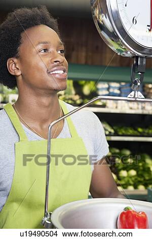 Picture - African American male store clerk weighing bell pepper. Fotosearch