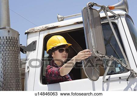 Stock Image - Asian female industrial worker adjusting mirror while sitting in logging truck. Fotosearch