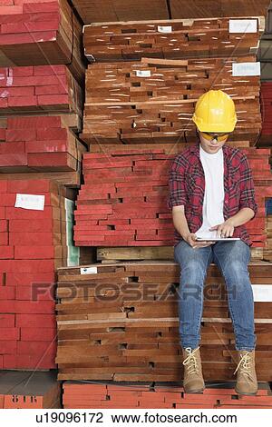 Stock Image - Asian female industrial worker using tablet PC while sitting on stacked wooden planks. Fotosearch
