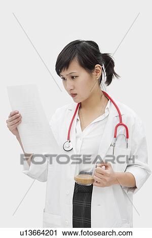 Stock Image - Asian female veterinarian reading document while having tea over gray background. Fotosearch