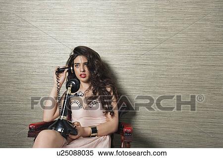 Stock Photography - Attractive young woman using nostalgic telephone while sitting on chair against textured background. Fotosearch