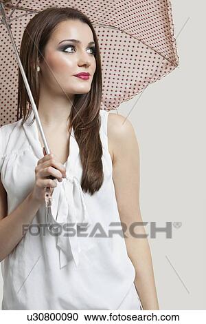 Beautiful young woman holding umbrella while looking away over gray background View Large Photo Image Stock Image - Beautiful young woman holding umbrella while looking away over gray background. Fotosearch