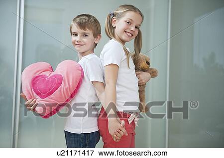 Stock Photo - Boy and girl with heart shape cushion and teddy bear holding hands while standing back to back. Fotosearch