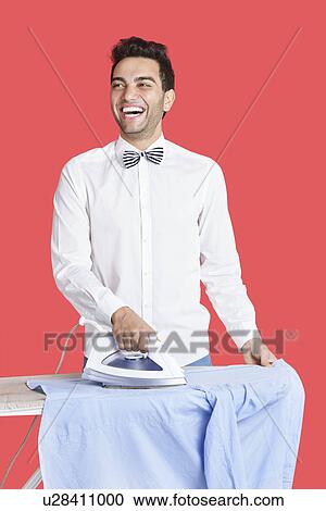 Stock Image - Cheerful man in formals ironing shirt over red background. Fotosearch