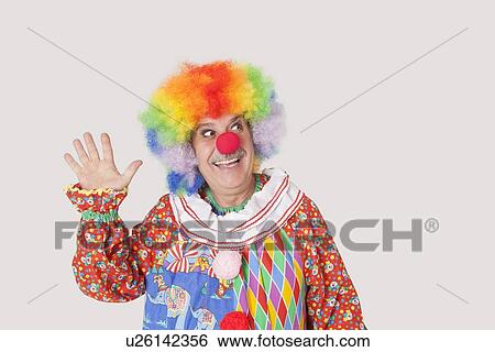 Stock Photograph - Cheerful senior male clown waving hand while looking away over gray background. Fotosearch