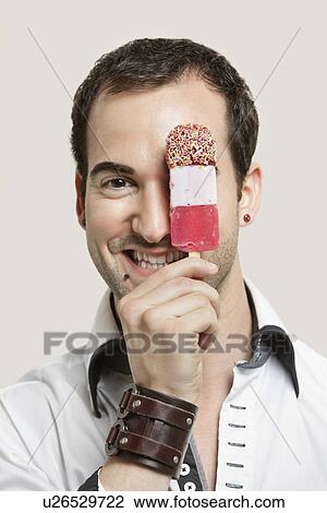 Stock Image - Close-up portrait of young man holding ice cream bar against gray background. Fotosearch
