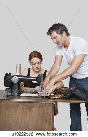 Stock Image - Designer helping coworker in stitching cloth on sewing machine over colored background. Fotosearch