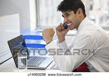 Stock Photo - Excited businessman using cell phone at office desk. Fotosearch