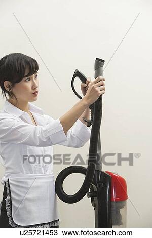 Stock Image - Female housekeeper holding vacuum cleaner pipe against gray background. Fotosearch