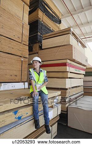 Stock Image - Female industrial worker having coffee while sitting on stack of wooden planks. Fotosearch
