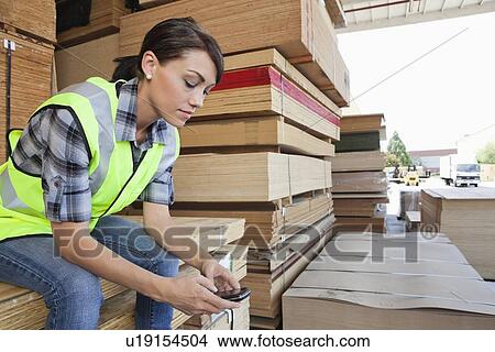 Picture - Female industrial worker using cell phone while sitting on stack of wooden planks. Fotosearch