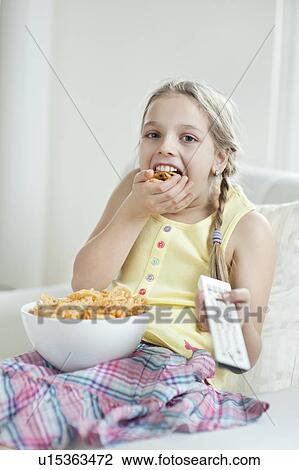 Stock Image - Girl watching TV as she stuffs her mouth with wheel shape snack pellets. Fotosearch