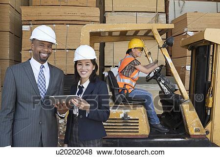 Stock Photo - Happy multiethnic engineers holding tablet PC with female worker driving forklift truck in background. Fotosearch