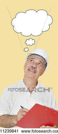 Stock Image - Happy senior constructor holding clipboard while looking up over yellow background. Fotosearch