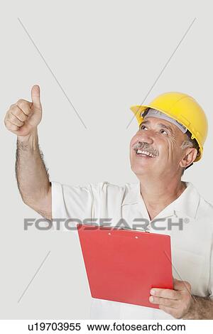 Stock Photography - Happy senior constructor holding clipboard while gesturing thumbs up over gray background. Fotosearch