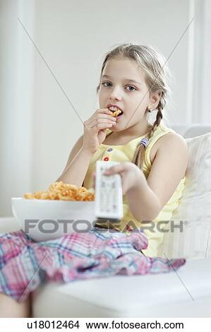Little girl watching TV as she eats wheel shape snack pellets View Large Photo Image Picture - Little girl watching TV as she eats wheel shape snack pellets. Fotosearch