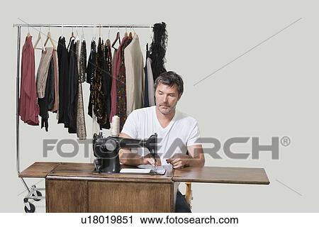 Stock Image - Mature male dressmaker sitting at sewing machine over colored background. Fotosearch