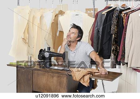 Stock Photograph - Mature male tailor thinking while sitting at sewing machine with fabric. Fotosearch
