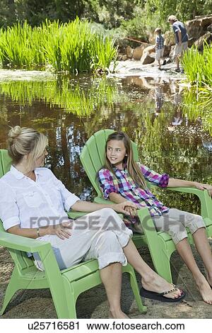 Stock Image - Mother and daughter relaxing on chairs with man and boy fishing in the background. Fotosearch