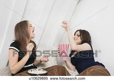 Stock Photography - Playful young female tossing popcorn into friend's mouth in bed. Fotosearch