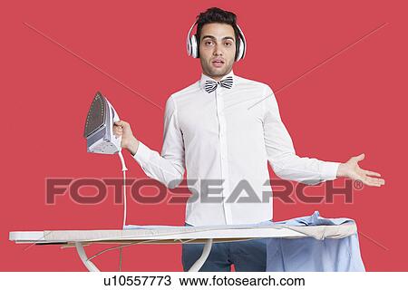Stock Image - Portrait of a confused man in formals ironing as he listens to music over red background. Fotosearch