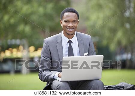 Stock Image - Portrait of a happy African American businessman using laptop in park. Fotosearch